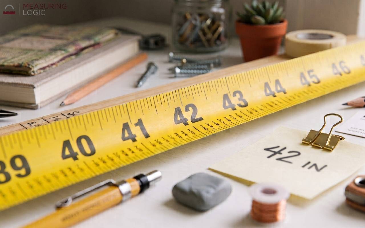 Close-up photograph of a yellow measuring tape resting on a white desk alongside various office supplies, with a shallow depth of field highlighting the 42-inch mark and a small clipped note reading "42 IN".