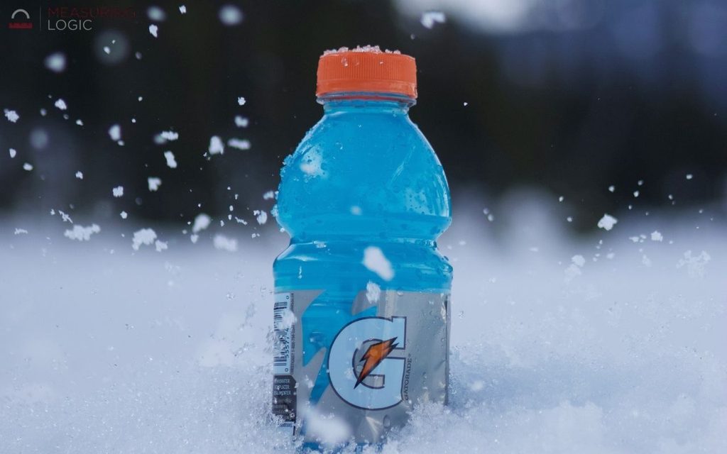 A close-up shot of a blue Gatorade bottle with an orange cap partially embedded in fresh snow while snowflakes drift around it.