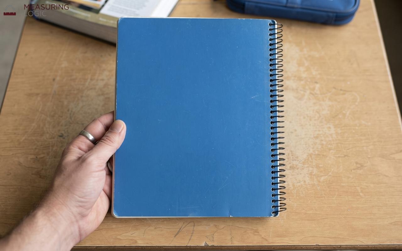 hand holding a blue spiral-bound notebook vertically against a wooden desk setup.